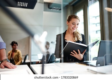 Beautiful Smiling Young Businesswoman Reading From Book While Standing At Desk Seen Through Glass Protection Shield At Office During COVID-19