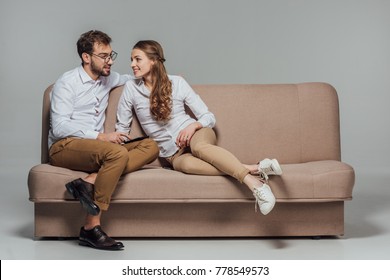 Beautiful Smiling Young Couple Using Smartphone Together While Sitting On Sofa Isolated On Grey 