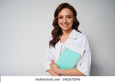 Beautiful smiling young female doctor in white medical jacket isolated on white background. Brunette woman cosmetologist holding books, notebook, folder. Space for text