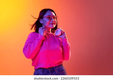 Beautiful smiling young girl in cute sweater  trendy sunglasses   headphones posing gradient orange background in neon light  Concept emotions  facial expression  youth  inspiration  sales  ad