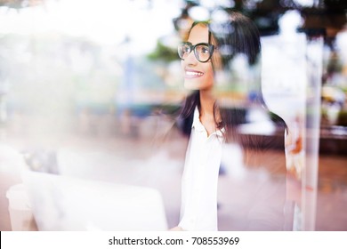 A Beautiful Smiling Young Professional Beautiful Indian Woman Wearing A Suit And Spectacles Working On A Laptop. View Through The Window 