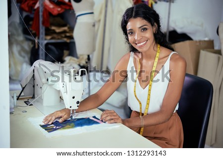 Beautiful smiling young seamstress working on sewing machine in workshop