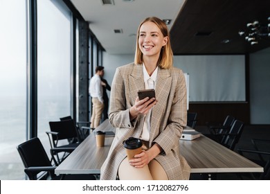 Beautiful smiling young smart businesswoman standing in the office with group of colleagues on the background, using mobile phone