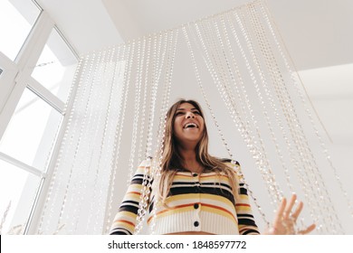 Beautiful Smiling Young Woman In Bright Living Room With White Walls And Iridescent Curtain