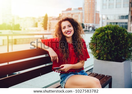 Beautiful smiling young woman with curly hair holds a mobile phone, spending time with pleasure while sitting on bench.