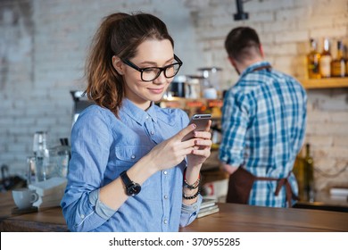 Beautiful smiling young woman in glasses standing in coffee shop and using mobile phone while barista working - Powered by Shutterstock