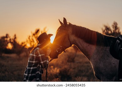 Beautiful smiling young woman in hat and shirt playing with brown horse in a field at sunset. Horseback riding. - Powered by Shutterstock