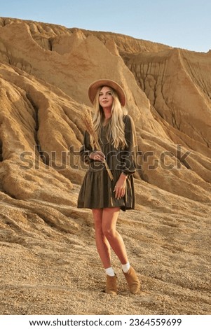 beautiful smiling young woman with long blonde hair in a hat against a sandy mountain background in sunset sunlight with a bouquet of dry grass in rustic style