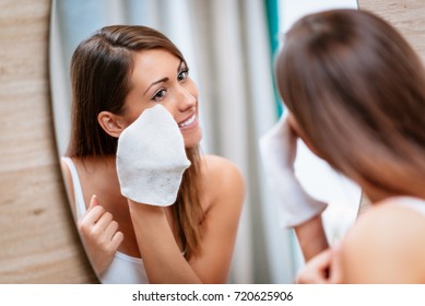 Beautiful smiling young woman removing make up in front of mirror.