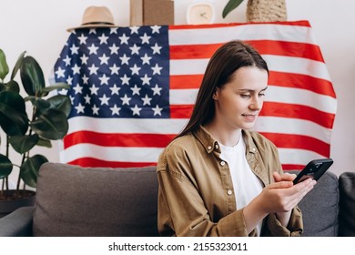 Beautiful smiling young woman sitting on cozy sofa at home with USA flag, cute happy caucasian female 20s old years with smartphone looking Independence Day celebration. Patriotic US holiday concept