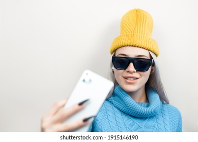 Beautiful Smiling Young Woman In Sunglasses Holding Smartphone, Indoors. Teenager Girl In Yellow Hat And Sweater Using Phone Camera. Concept Story, Social Media, Selfie, Blogging.