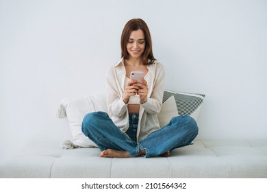 Beautiful smiling young woman teenager girl student with dark long hair in white shirt using mobile phone in hand sitting on couch at home - Powered by Shutterstock