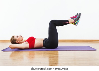 Beautiful sportive girl wearing snickers, black leggings and red short top doing abdominal exercise on purple matt at gym, fitness, white wall and wooden floor, copy space.