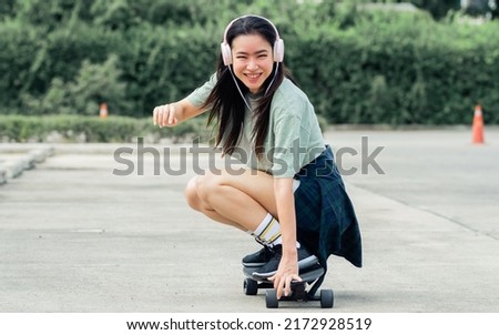 Beautiful sportive hipster happy Asian woman wearing casual shirt and shorts with headphone to listen music while playing skateboard as hobby in free time day on holiday, smiling at outdoor park