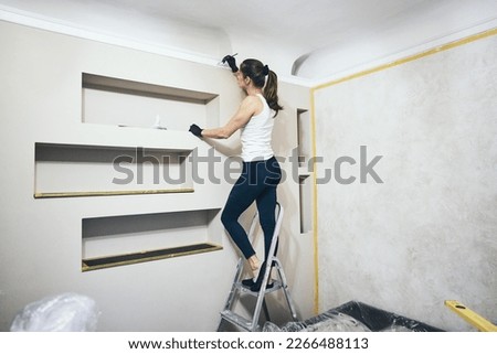 A beautiful sportive slim girl in a white tank top and black latex gloves paints the wall in the room with a brush standing on a ladder. Renovation and repair concept.