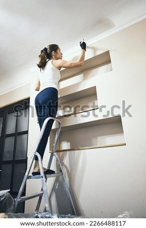 A beautiful sportive slim girl in a white tank top and black latex gloves paints the wall in the room with a brush standing on a ladder. Renovation and repair concept. Vertical shot.