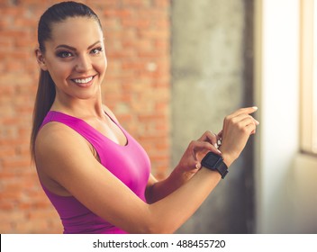 Beautiful sports girl is touching her fitbit, looking at camera and smiling while standing in fitness hall