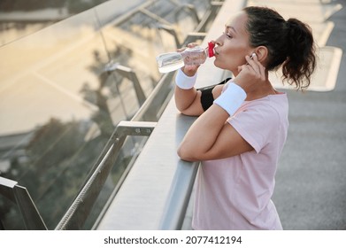 Beautiful sportswoman drinks water from bottle to hydrate her body after outdoor morning cardio workout. Young sportive woman resting after jogging on the city bridge against the background of nature - Powered by Shutterstock