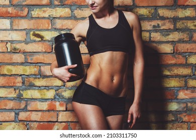 Beautiful Sportswoman Looking Down And Holding A Jar Of Protein, Standing Against Brick Wall, Cropped