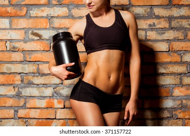 Beautiful Sportswoman Looking Down And Holding A Jar Of Protein, Standing Against Brick Wall, Cropped