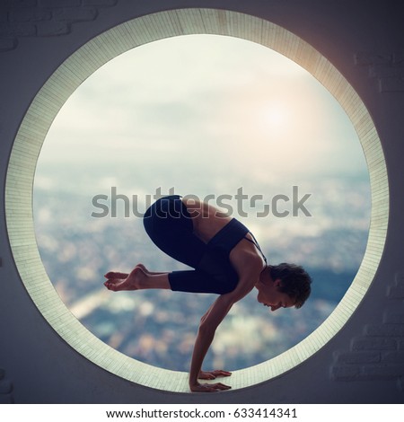 Beautiful sporty fit yogi woman practices yoga asana Bakasana - crane pose in a round window with a view of the city at sunset
