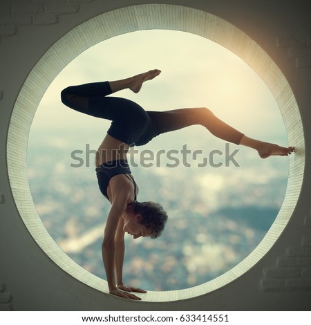 Beautiful sporty fit yogi woman practices yoga handstand asana Bhuja Vrischikasana - Scorpion handstand pose in a round window with a view of the city at sunset