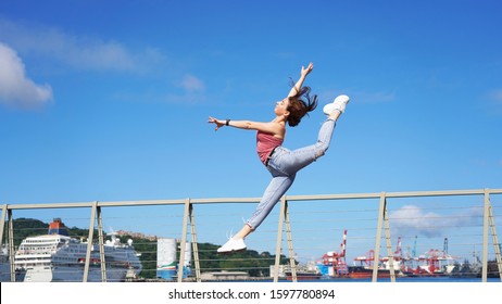 beautiful sporty girl in life style clothes jumping dance pose on the observation deck on a sunny day. circus gymnast in the port in denim casual wear. acrobatic twine jumping. gymnastic split in jump