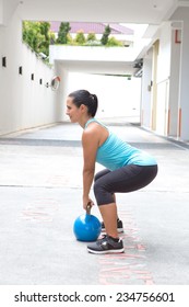 Beautiful Sporty Hispanic Woman In Blue Attire Holding A Blue Kettlebell In Dead Lift Post Outdoors