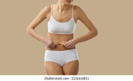 Beautiful sporty perfect skinny Caucasian woman in underwear, standing on beige background, pinches skin on naked belly, unsatisfied with diet result, worrying about body fat, thinking she's imperfect