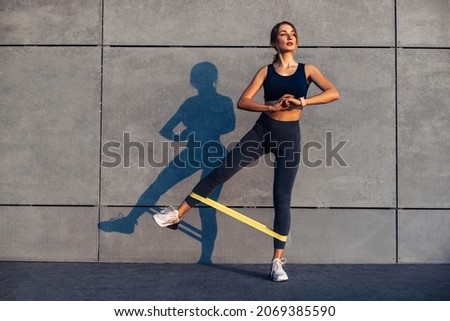 Beautiful sporty woman doing exercises with resistance band, fitness woman doing leg exercises with yellow fitness rubber band on gray background