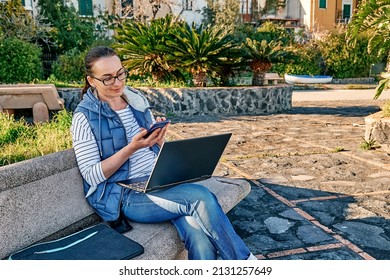 Beautiful sporty woman wearing eyeglasses with laptop computer working outdoors while sitting on bench at the seaside. Modern lifestyle, connection, blogging, business, freelance work concept.