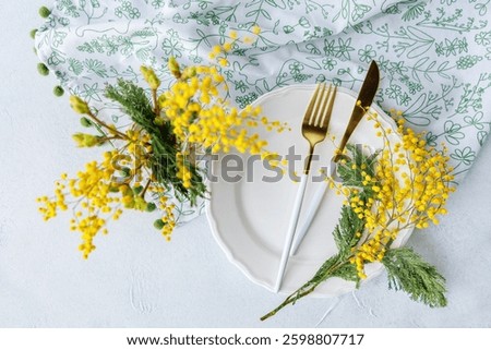 Beautiful spring table setting with white plate, golden cutlery, and fresh mimosa flowers on a light background with a floral napkin.