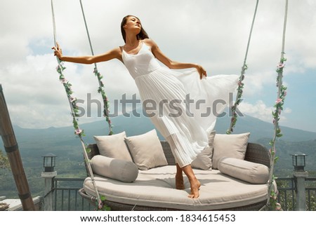 Beautiful Girl’s Standing On Sky Swing Portrait In Bali, Indonesia. Young Woman In White Dress Fluttering In Wind Posing On Hanging Chair With Flowers Against Mountain. 