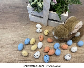 Beautiful Still Life. Many Colorful Small Chocolate Eggs On Wooden Background. Green Leaves With Pink Flowers And Bird In Wooden Basket. Place For Inscription. Preparation For Easter. Bunny Plush Toy