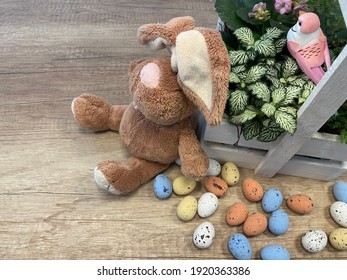 Beautiful Still Life. Many Colorful Small Chocolate Eggs On Wooden Background. Green Leaves With Pink Flowers And Bird In Wooden Basket. Place For Inscription. Preparation For Easter. Bunny Plush Toy