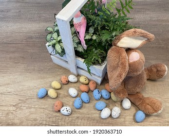 
Beautiful Still Life. Many Colorful Small Chocolate Eggs On Wooden Background. Green Leaves With Pink Flowers And Bird In Wooden Basket. Place For Inscription. Preparation For Easter. Bunny Plush Toy