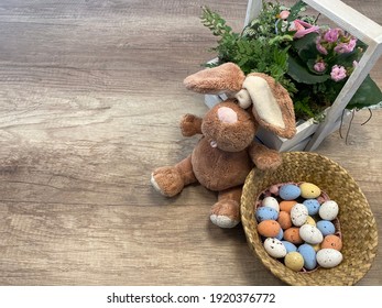 Beautiful Still Life, Wooden Background. Braided Basket With Small Chocolate Eggs. Green Leaves Of Plants, Pink Flower And Bird In Wooden Space. Copy Place. Preparation For Easter. Bunny Plush Toy