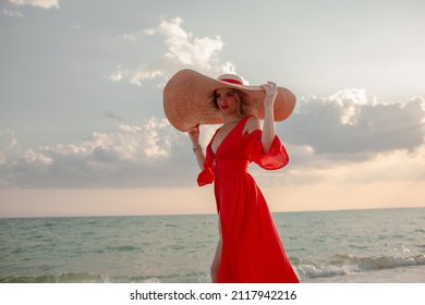Beautiful strong woman wearing maxi straw hat and red chiffon cape like dresss walking in the beach against the blue sky and sea