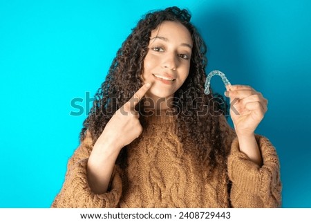 Beautiful student girl holding books wearing formal clothes holding an invisible aligner and pointing perfect straight teeth. Dental healthcare and confidence concept.