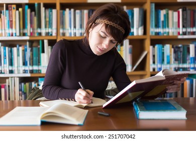 Beautiful student girl studying in college library. Writing notes from book at her notebook. Selective focus on face. 