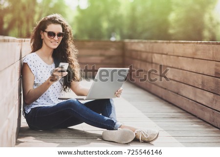Beautiful student girl working on laptop outdoor. Young woman using smartphone and laptopcomputer in the park