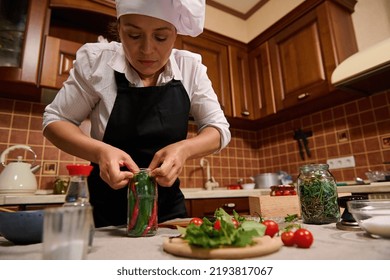 Beautiful Stunning Woman, Housewife Wearing White Chef's Cap And Black Kitchen Apron Inserting Red And Green Hot Chili In A Sterilized Glass Jar, Preparing Fermented Delicacies And Marinated Food