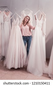 Beautiful style. Joyful young woman standing near the wedding dresses while choosing between them