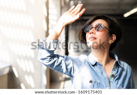 Beautiful / Stylish Asian woman wearing 100% UV light eyes protection sunglasses, stand and raise her hand to block out bright glare and sunlight from outside to avoid ultraviolet rays over exposure. 