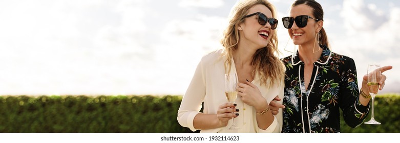 Beautiful and stylish friends with champagne laughing outdoors. Two young women wearing sunglasses holding wine glasses with lots of copy space. - Shutterstock ID 1932114623