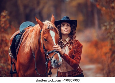 Beautiful stylish girl dressed as an American cowboy is on a rural road with a saddled horse, golden autumn