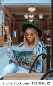 Beautiful stylish girl in jeans jacket and hat is reading a newspaper while resting in a cafe outdoors. Portrait of cute girl sitting on the wooden bench with open newspaper