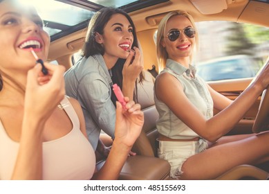 Beautiful Stylish Girls Are Smiling And Having Fun While Driving The Car. Two Girls Are Using Lipstick, Attractive Driver Is Looking At Camera