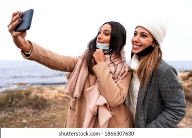 Beautiful Stylish Hispanic Woman Making Selfie Outdoor In Sea Winter Vacation With Her Caucasian Girlfriend Wearing Lowered Protection Mask - Young Female LGBTQ Couple Using Phone For A Self Portrait