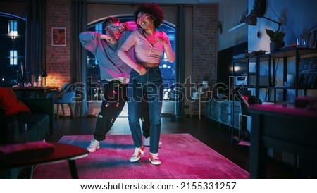 Beautiful Stylish Multiethnic Couple in Casual Outfits and Futuristic Neon Glowing Glasses, Dance and Have a Party at Home in Loft Apartment. Recording Funny Viral and Active Videos for Social Media. 商業照片 © 